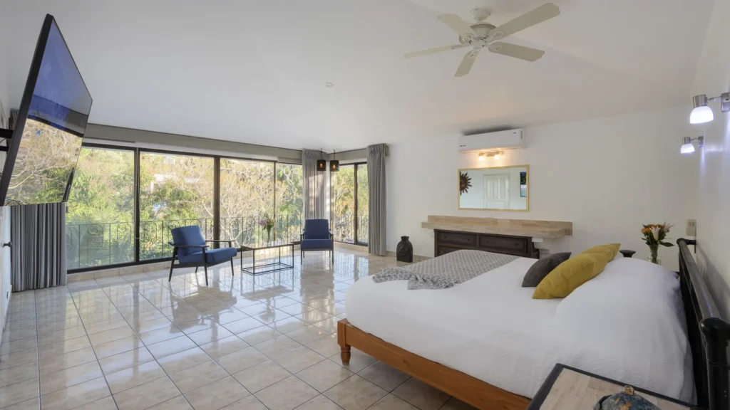 Bright hotel room with a large glass wall and balcony, two blue chairs, small glass table, and a king bed on the right.