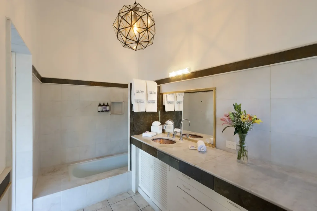 Bright marble bathroom with a double-sink vanity, gold-framed mirror, towels, and a geometric pendant light overhead. A vase of flowers sits on the counter beside the sinks.