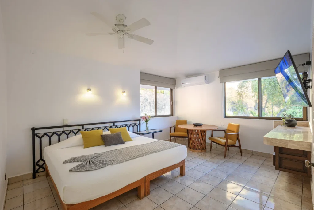 Bright hotel room with a white bed and yellow-gray accent pillows, a round wooden dining table with two mustard chairs, and a wall-mounted TV by large windows.