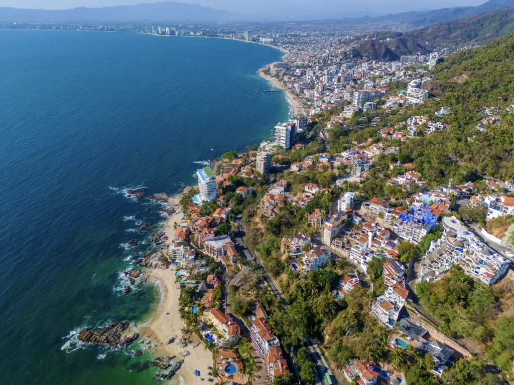 Aerial view of a coastal city along a curved shoreline, with high-rise buildings and houses on a green hillside above the blue sea.