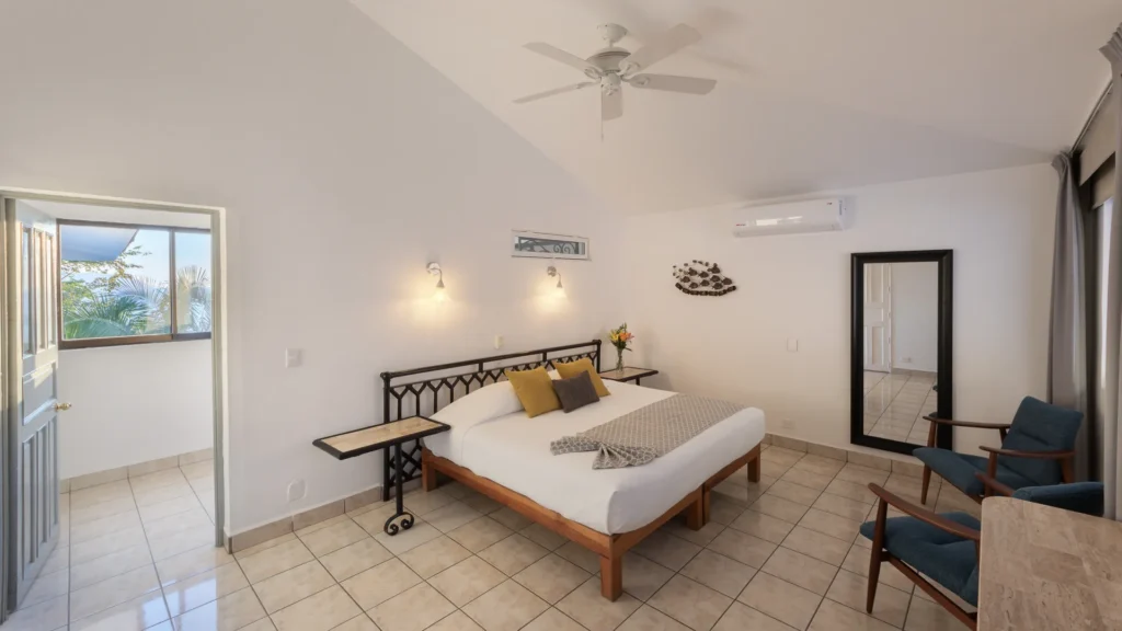 Bright hotel bedroom with a metal-framed bed, two bedside tables with lamps, and a ceiling fan above. Nearby is a doorway to a sunny balcony with palm trees outside.