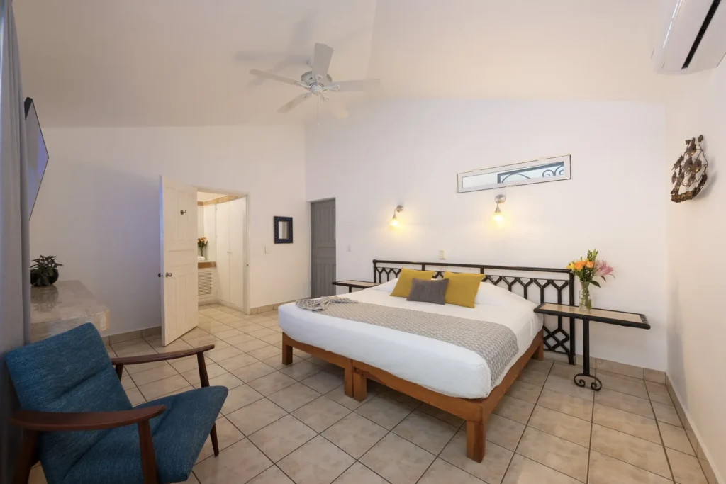 Bright hotel room with a large bed, yellow and gray pillows, and nightstands on either side; vase of flowers on one table.