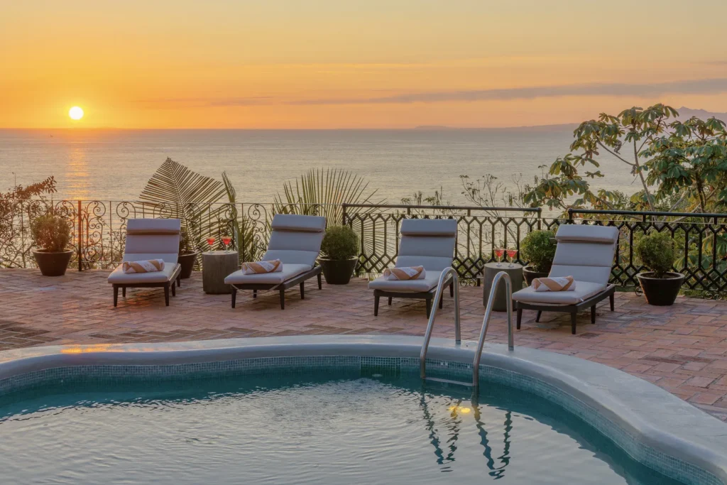 Poolside terrace at sunset with four cushioned lounge chairs overlooking the sea and a decorative railing.