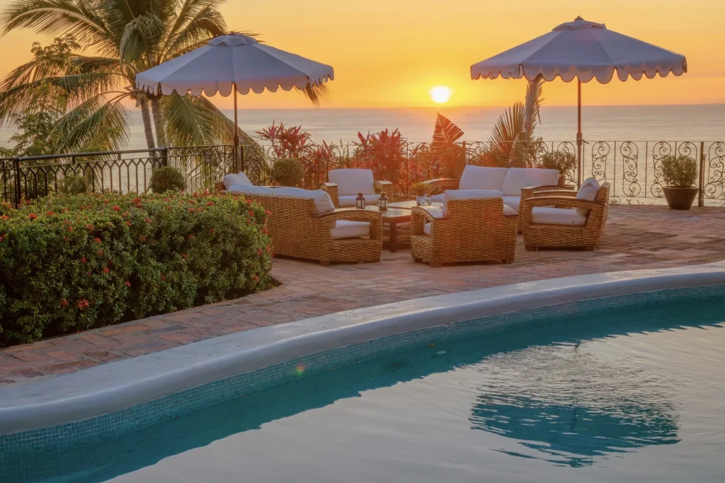 Poolside patio at sunset with wicker chairs and white cushions, two umbrellas, and tropical plants overlooking the ocean.