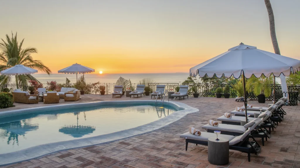 Poolside at a tropical resort during sunset with white umbrellas, lounge chairs, and palm trees overlooking the ocean far horizon.