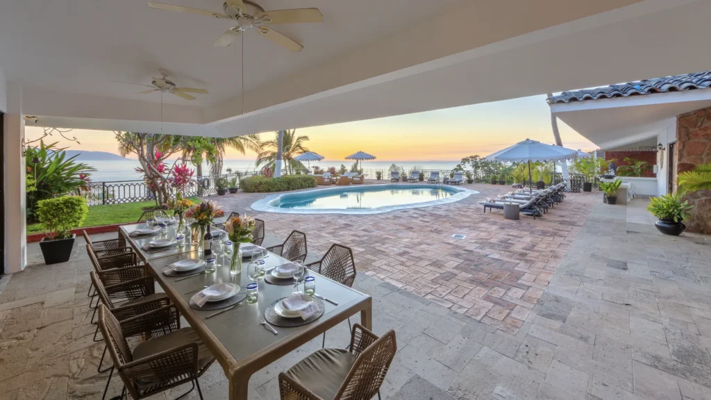 Outdoor dining area overlooking a curved pool and ocean horizon at sunset, table set with plates, glasses, and flowers ready for a meal.