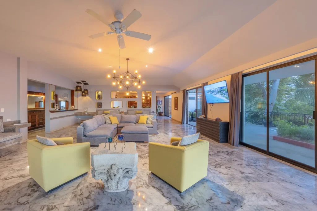Open-concept living room with marble floors, a gray sectional sofa, yellow armchairs, a modern chandelier, and large sliding glass doors to an outdoor patio.