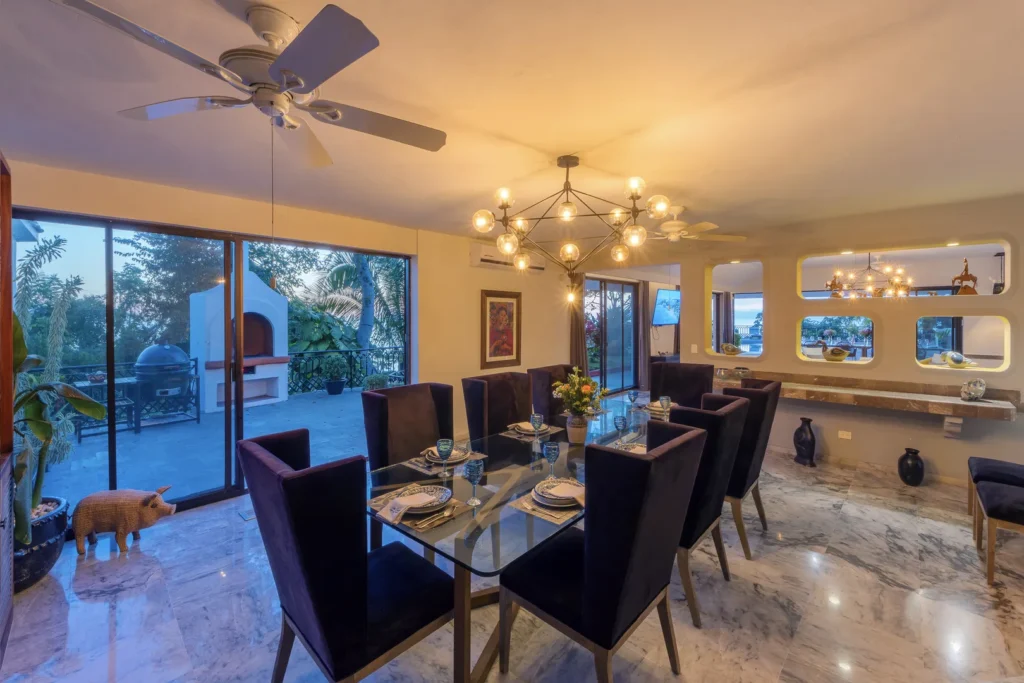 Luxurious dining room with a glass-top table set for eight, purple velvet chairs, and a geometric chandelier, large glass doors open to a leafy outdoor patio.