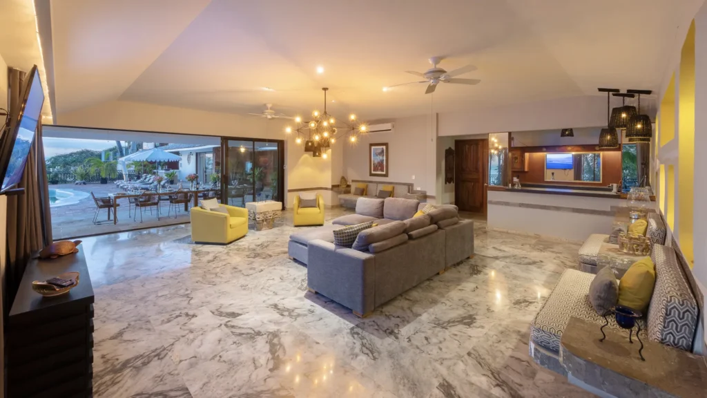 Spacious living room with marble floors, gray sectional, yellow chairs, and a chandelier, opening to an outdoor dining area.