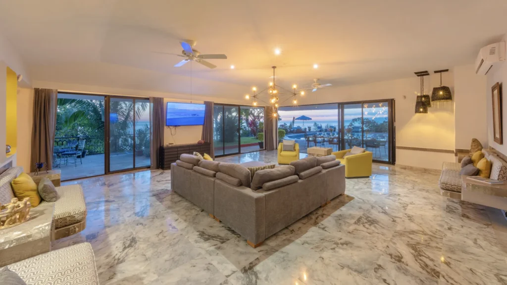 Open-plan living room with marble floor, gray sectional sofa, yellow chairs, and large glass doors to a sunset ocean-view patio.