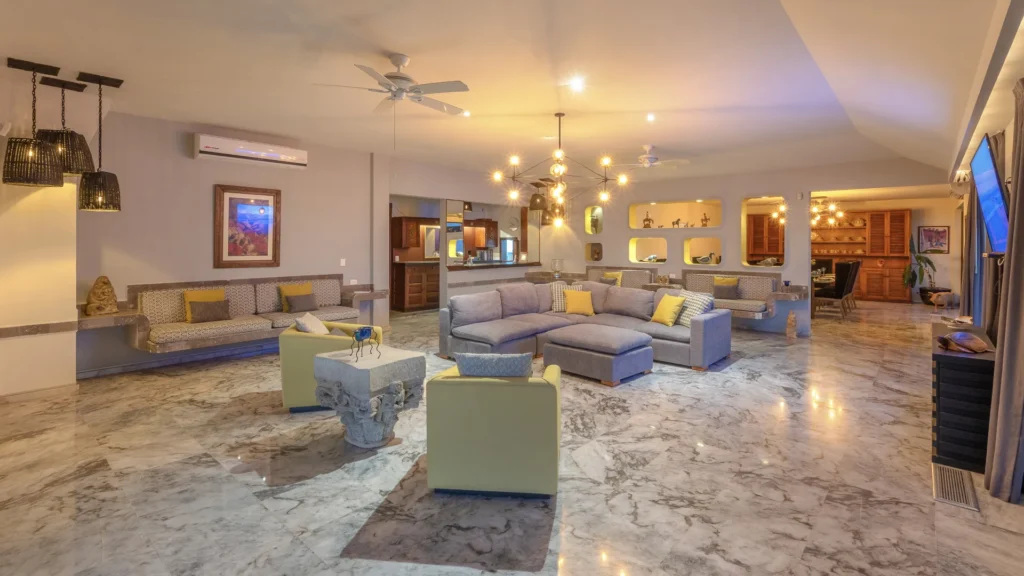 Spacious living room with marble floors, gray sectional sofa, and yellow cushions under warm pendant lights and ceiling fans.