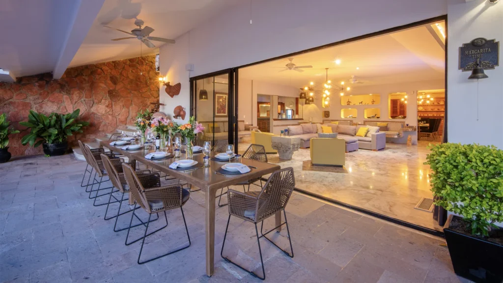 Outdoor dining area with a long wooden table set for eight, wicker chairs, flowers, and a stone wall leading into a bright living room.