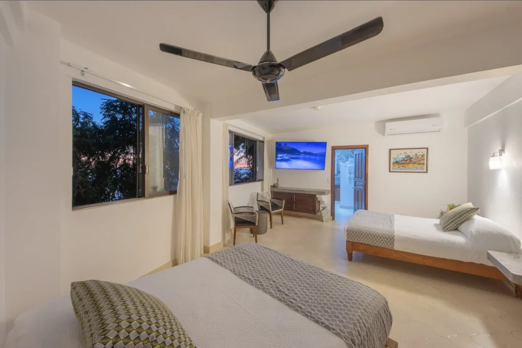 Bright hotel room with two queen beds, gray patterned runners, and matching pillows; ceiling fan overhead.