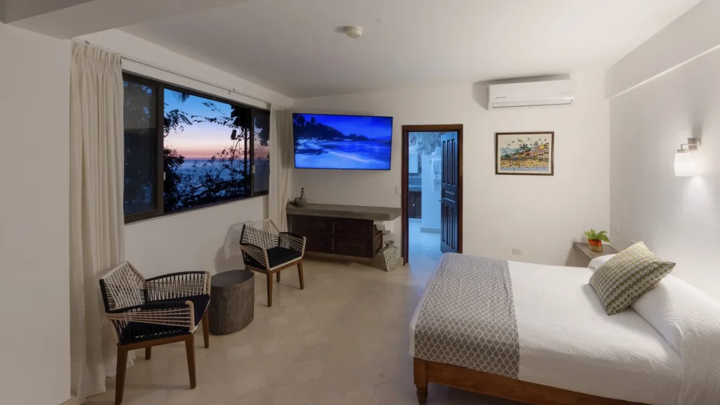 Hotel room with a large bed, dresser, wall TV, and a doorway to a bright bathroom area; sunset visible through the window outside the curtains.