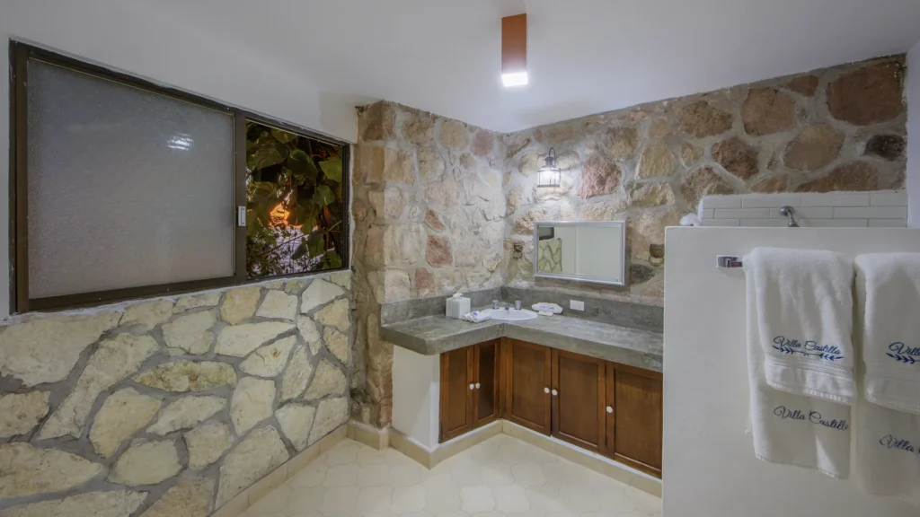 Bathroom with stone walls, wooden vanity, and a sink; fluffy white towels hang on a partition wall to the right.