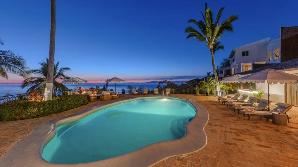 Poolside at a tropical resort during blue hour, with palm trees, lounge chairs, and a sea horizon in the background.