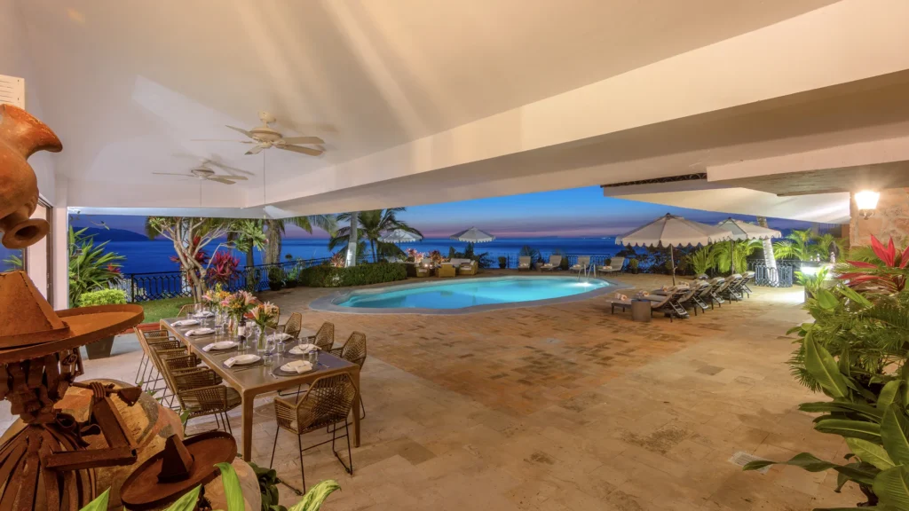 Poolside dining area with a long table set for a meal, chairs, and tropical plants under a covered patio overlooking an ocean sunset.