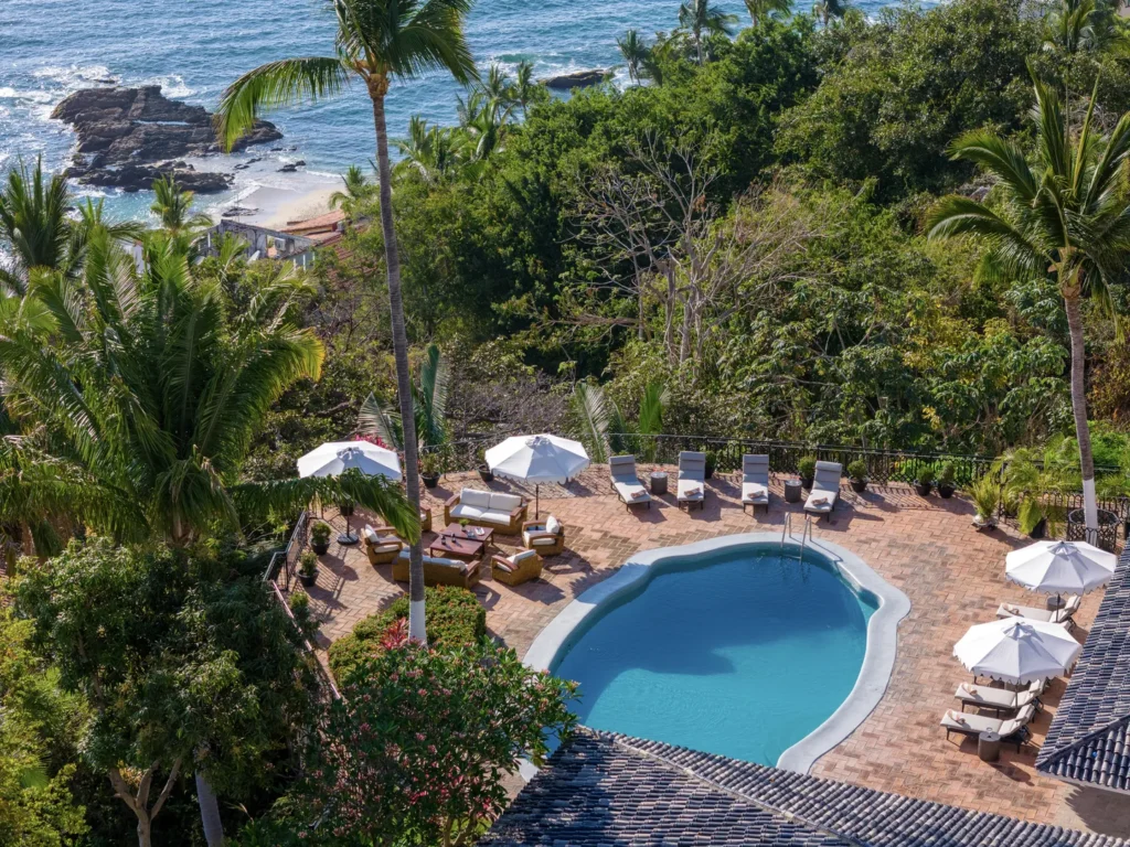 Overhead view of a tropical resort pool area with white lounge chairs, umbrellas, and a brick-patio surrounding the pool by the sea at the edge of lush greenery.