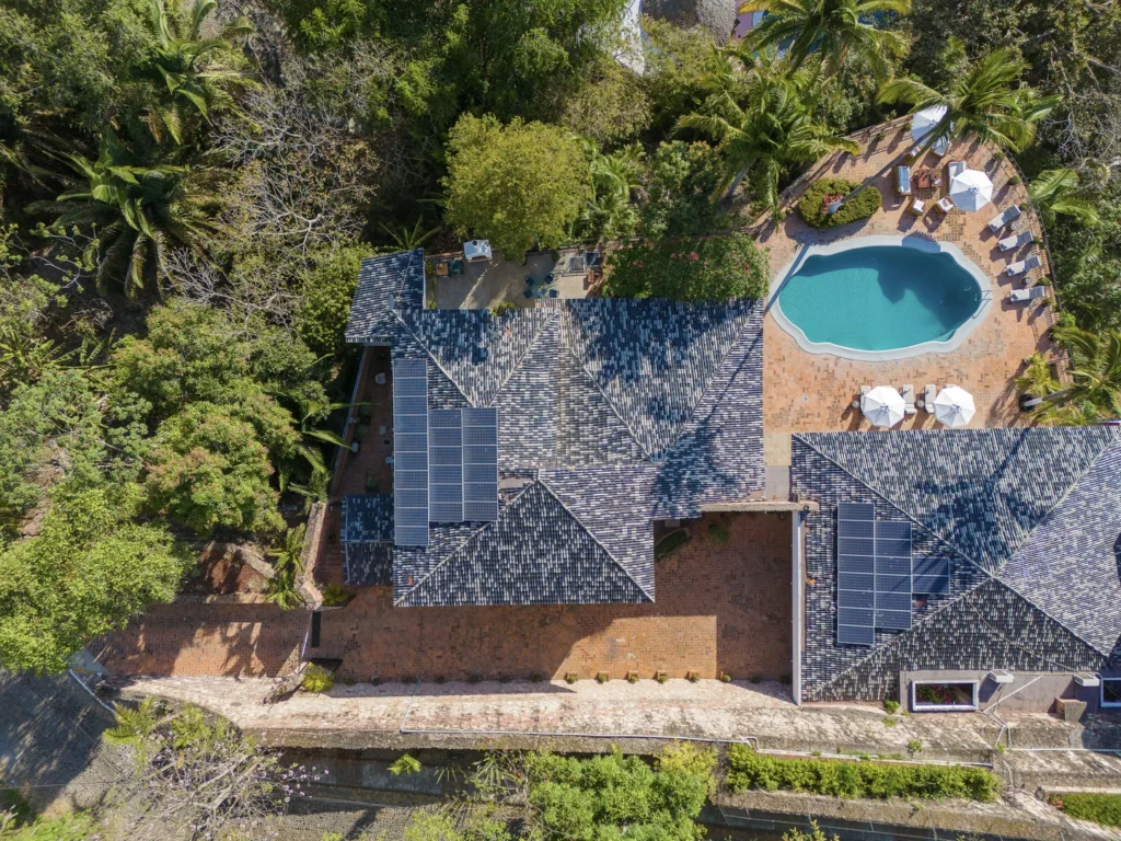 Aerial view of a house with solar panels on the roof, a kidney-shaped pool, and a brick patio surrounded by trees and greenery.