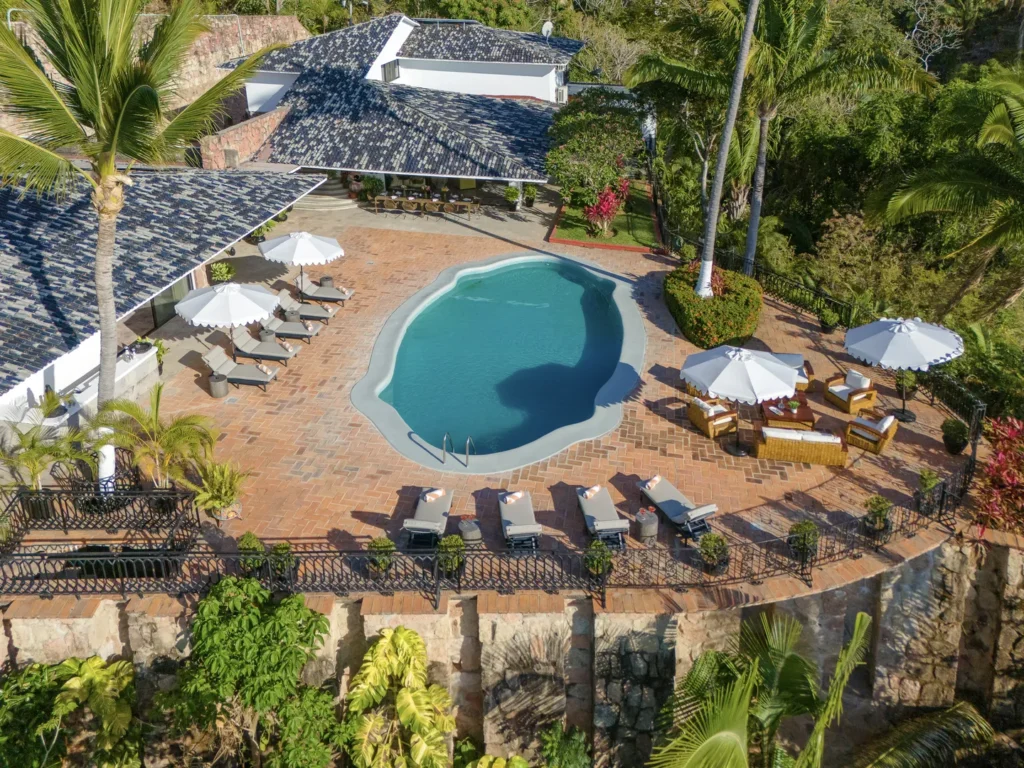 Aerial view of a resort pool area with a curved blue pool, terracotta deck, and white umbrellas surrounded by lounge chairs and palm trees.