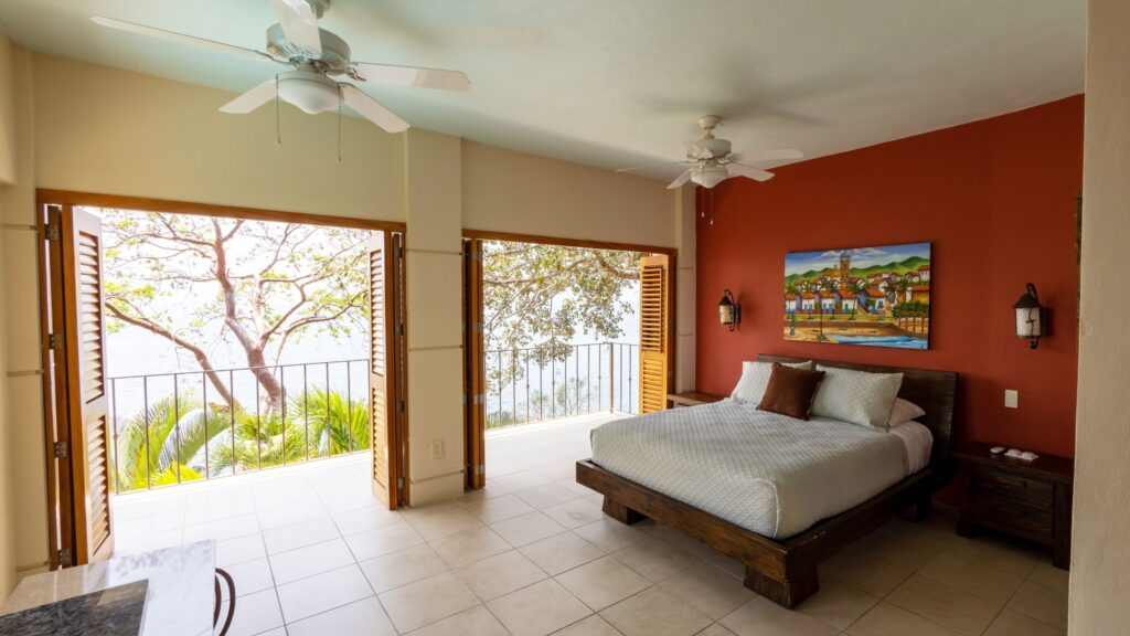 Hotel-style bedroom with a low wooden bed, white bedding, and a red accent wall displaying a tropical painting; sliding doors open to a balcony with an ocean view and trees outside.
