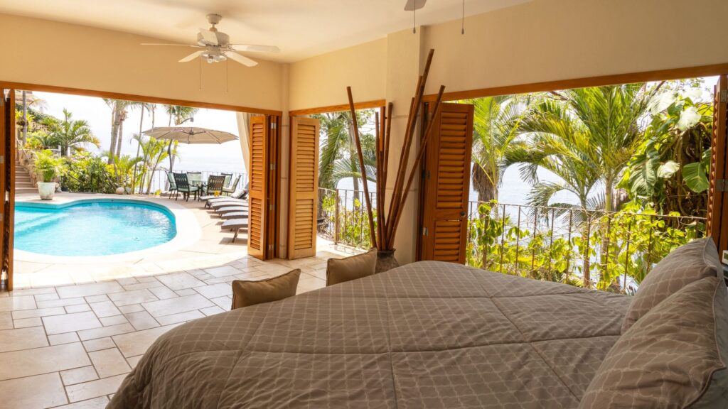 Bedroom with two queen beds opens to a poolside terrace; see ocean beyond, palm trees, and outdoor seating under umbrellas on a sunny day.