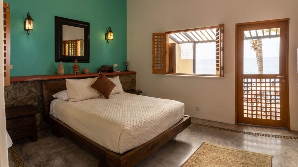 Bedroom with teal accent wall, white quilted bed, and a door opening to an ocean-facing balcony.