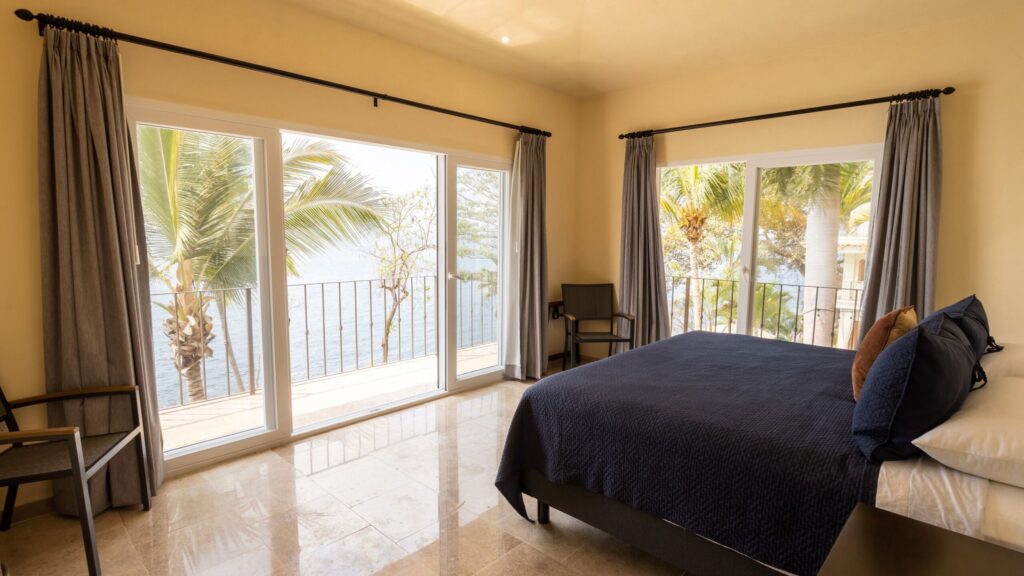 Bright hotel bedroom with navy bedspread, gray curtains, and large glass doors opening to a balcony with palm trees and water view.