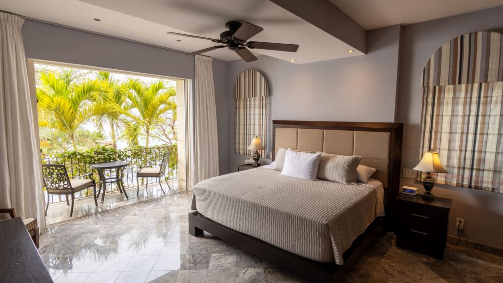 Hotel room with a large bed and balcony access, green palm trees visible outside the glass doors with a small table and chairs on the balcony