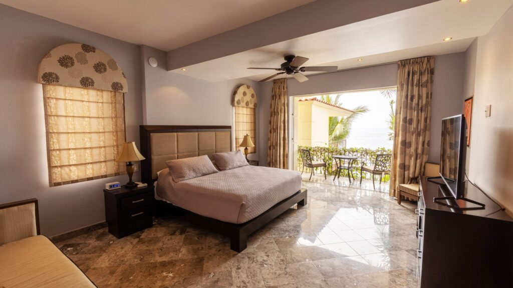 Hotel bedroom with a large bed, nightstands, lamps, and a ceiling fan, opening to a balcony with a sea view and palm trees outside