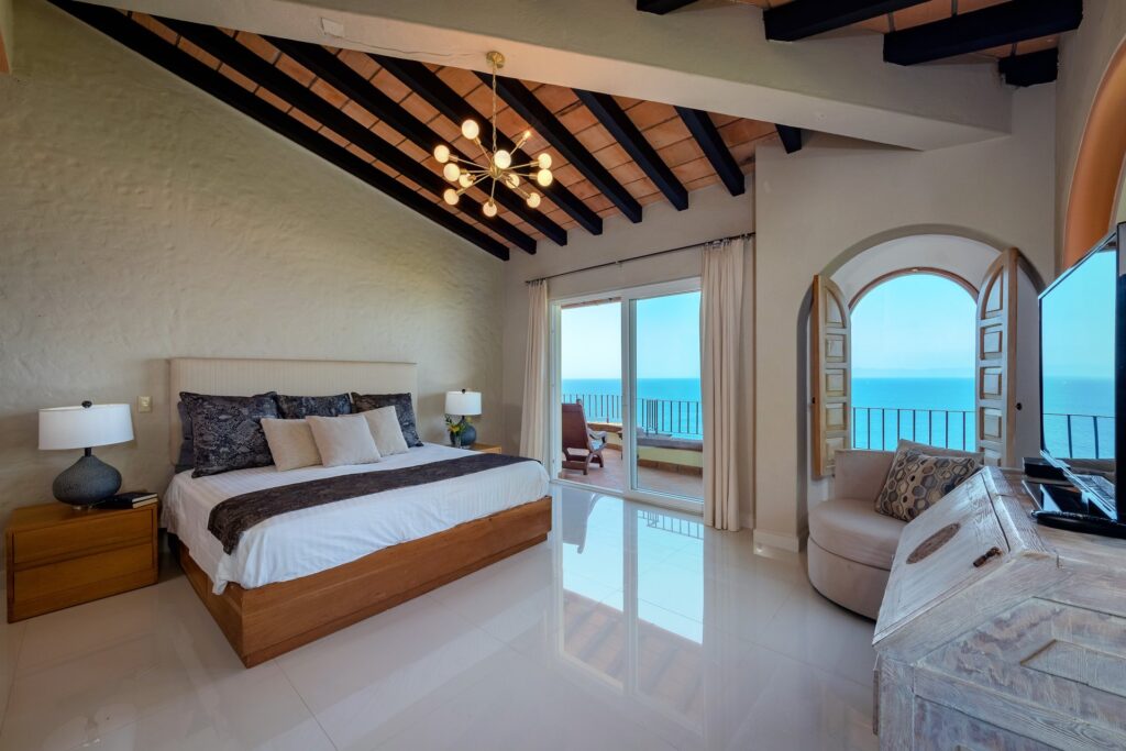 Bright bedroom with a wooden bed, matching nightstands, and a chandelier, opening to a balcony with an ocean view.